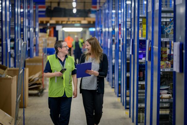 Warehouse staff discussing inventory management with shelves stocked with products in the background.
